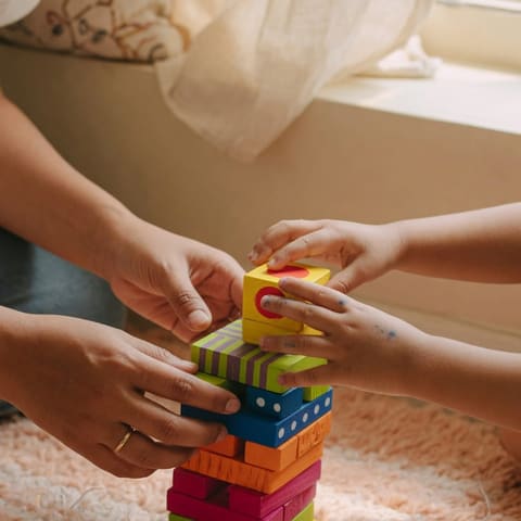 Close-up of a parent's hands and a young child's hands stacking colorful wooden blocks together on a soft rug