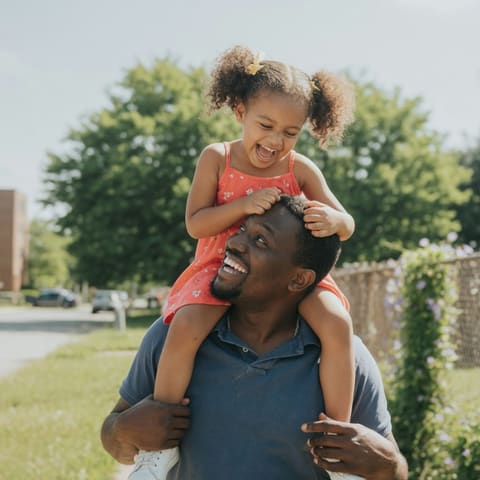 A father carrying his laughing young child on his shoulders outdoors on a bright sunny day